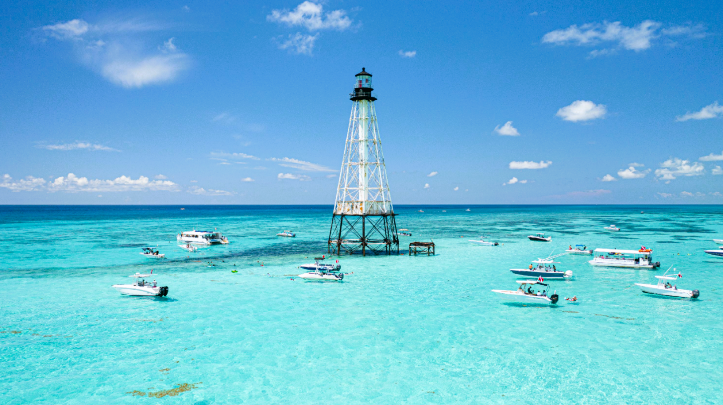 Turquoise sea with boats near a tall lighthouse under a clear blue sky.