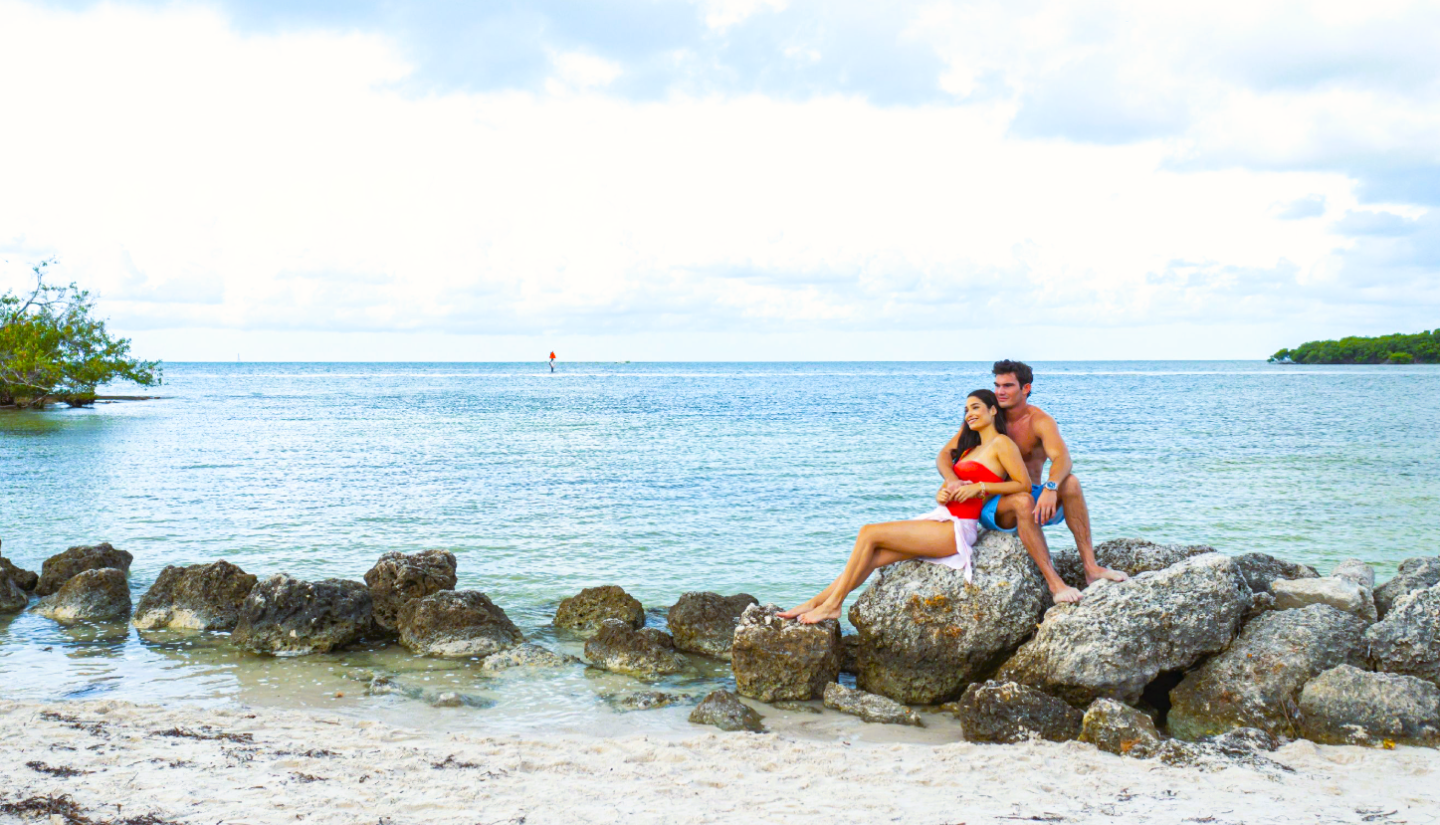 A couple sits on rocks by a calm sea, with trees in the background.