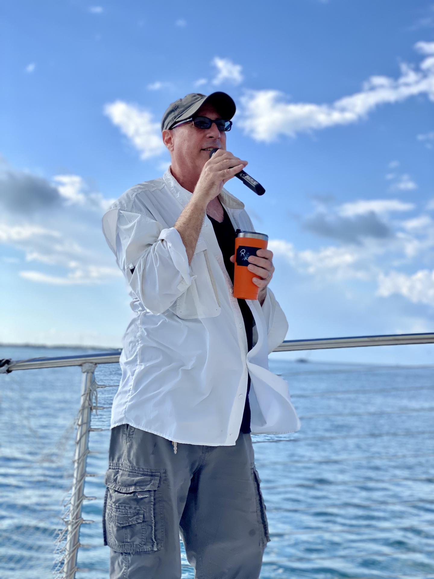 Man speaking into a microphone on a boat, holding an orange cup.