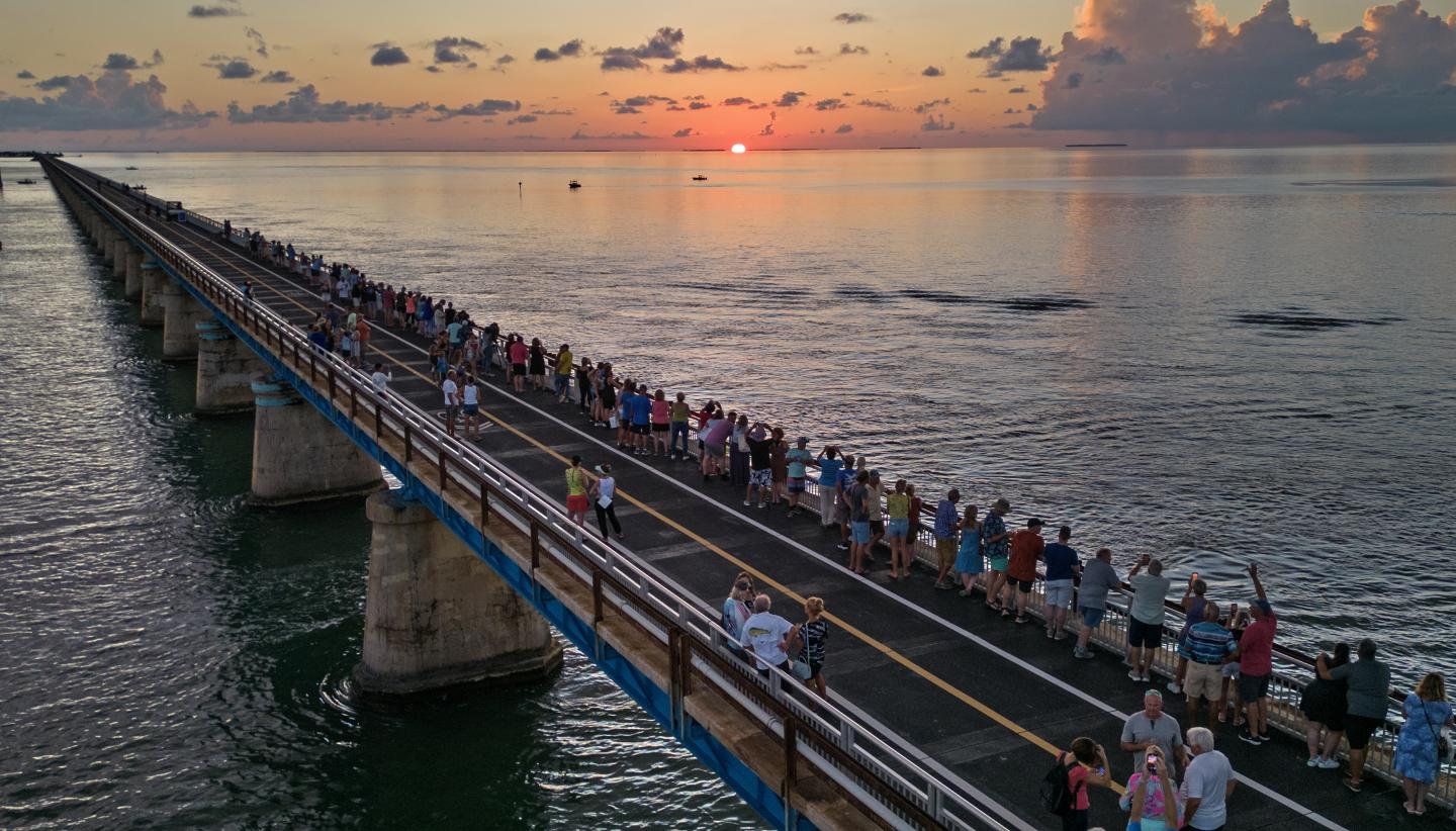 People walking on a bridge over the ocean at sunset.