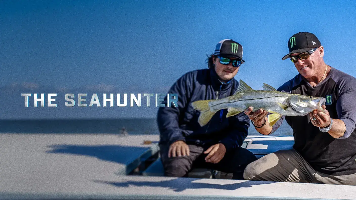 Two men on a boat, one holds a large fish. "The Seahunter" text appears.