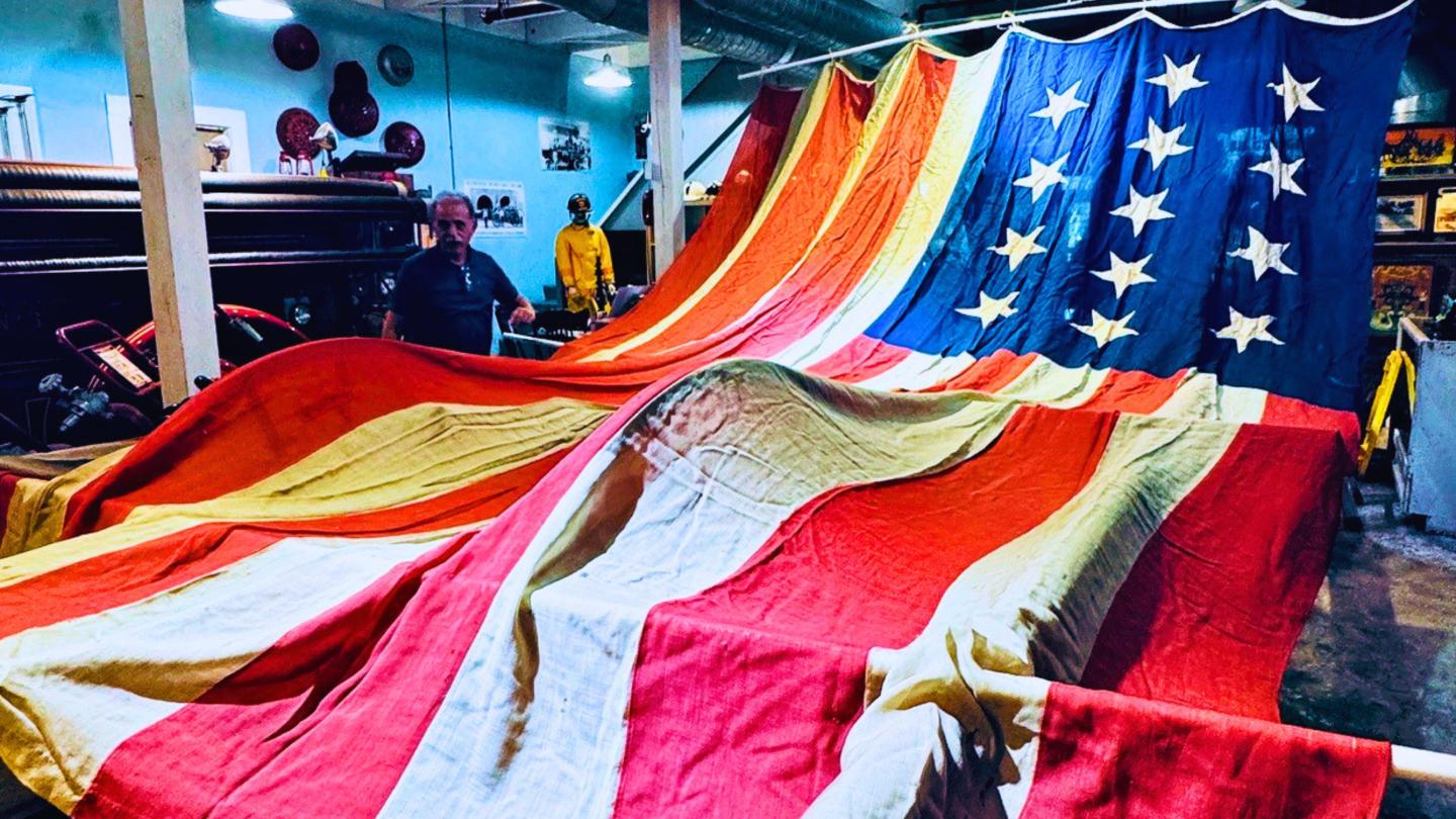 Large American flag with stars spread across a workshop floor with people observing.
