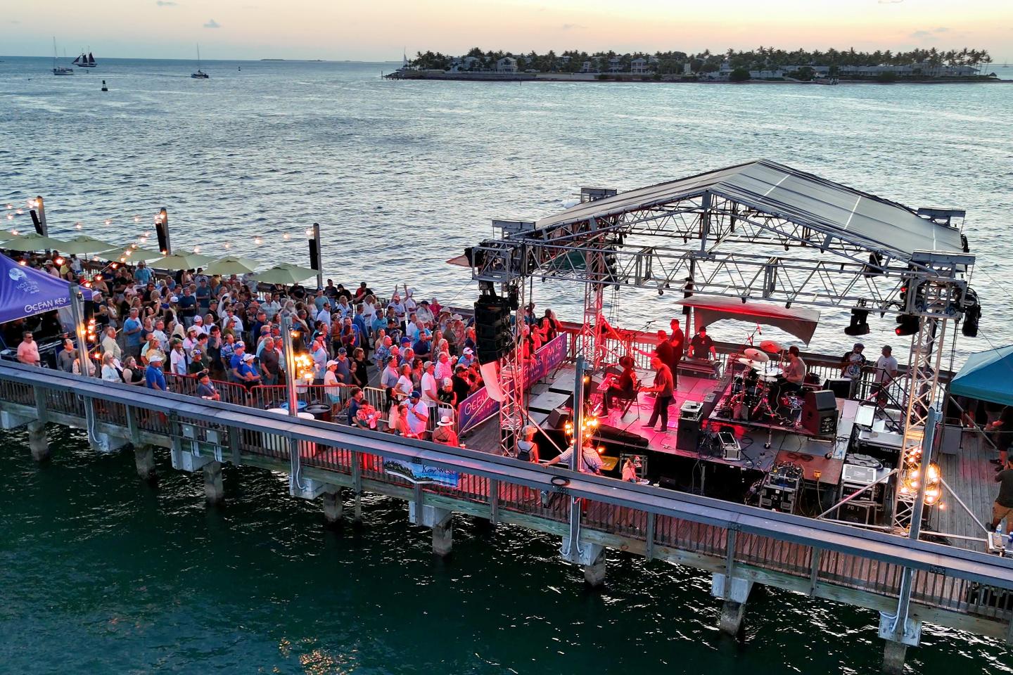 Concert on a pier at sunset, with a lively crowd and band under colorful lights.