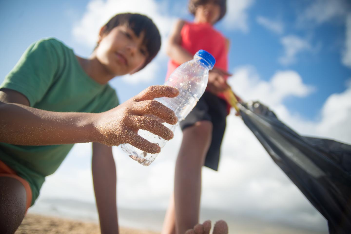 Child holding plastic bottle on a beach, another child with a trash bag in the background.