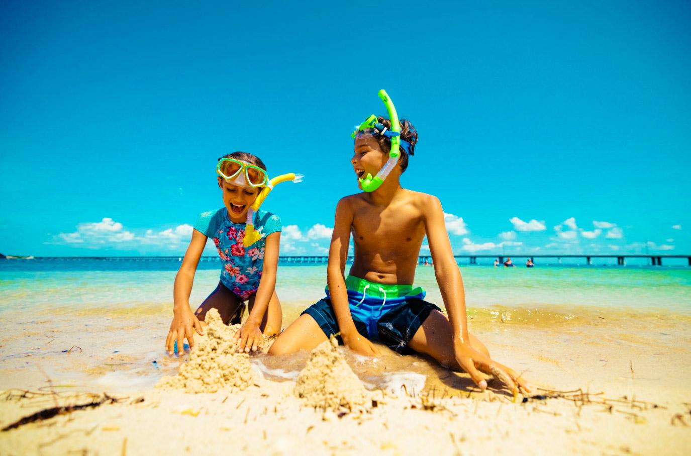 Two kids in snorkel gear play in the sand by the ocean.