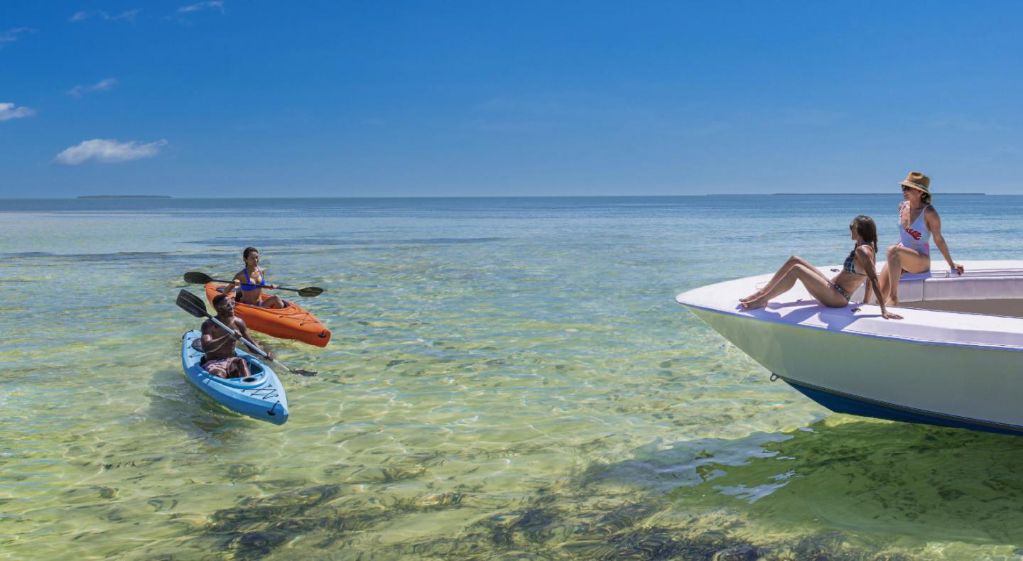 Two people kayaking, approaching a boat with two people relaxing, under a clear blue sky.