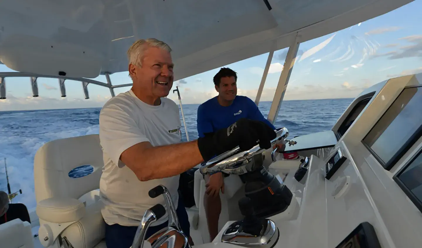 Elderly man smiling while steering a boat at sea, with another man seated nearby.