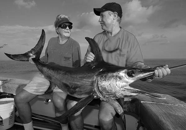 Two men on a boat holding a large swordfish, smiling.
