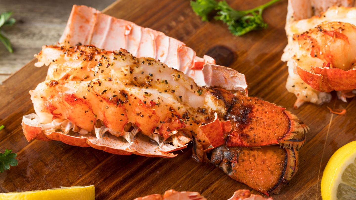 Grilled lobster tail on wooden board with herbs and lemon slice.