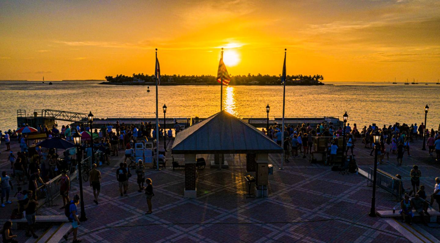 Crowd gathers at waterfront plaza during sunset.