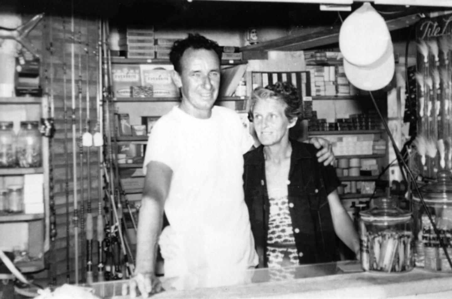 Man and woman smiling behind a store counter, surrounded by shelves.