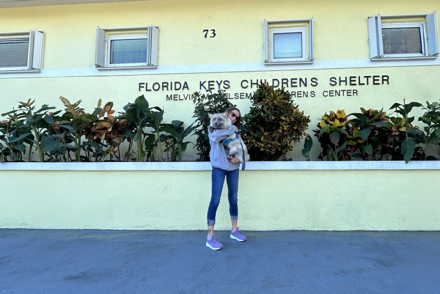 Woman holds child outside colorful building with plants.