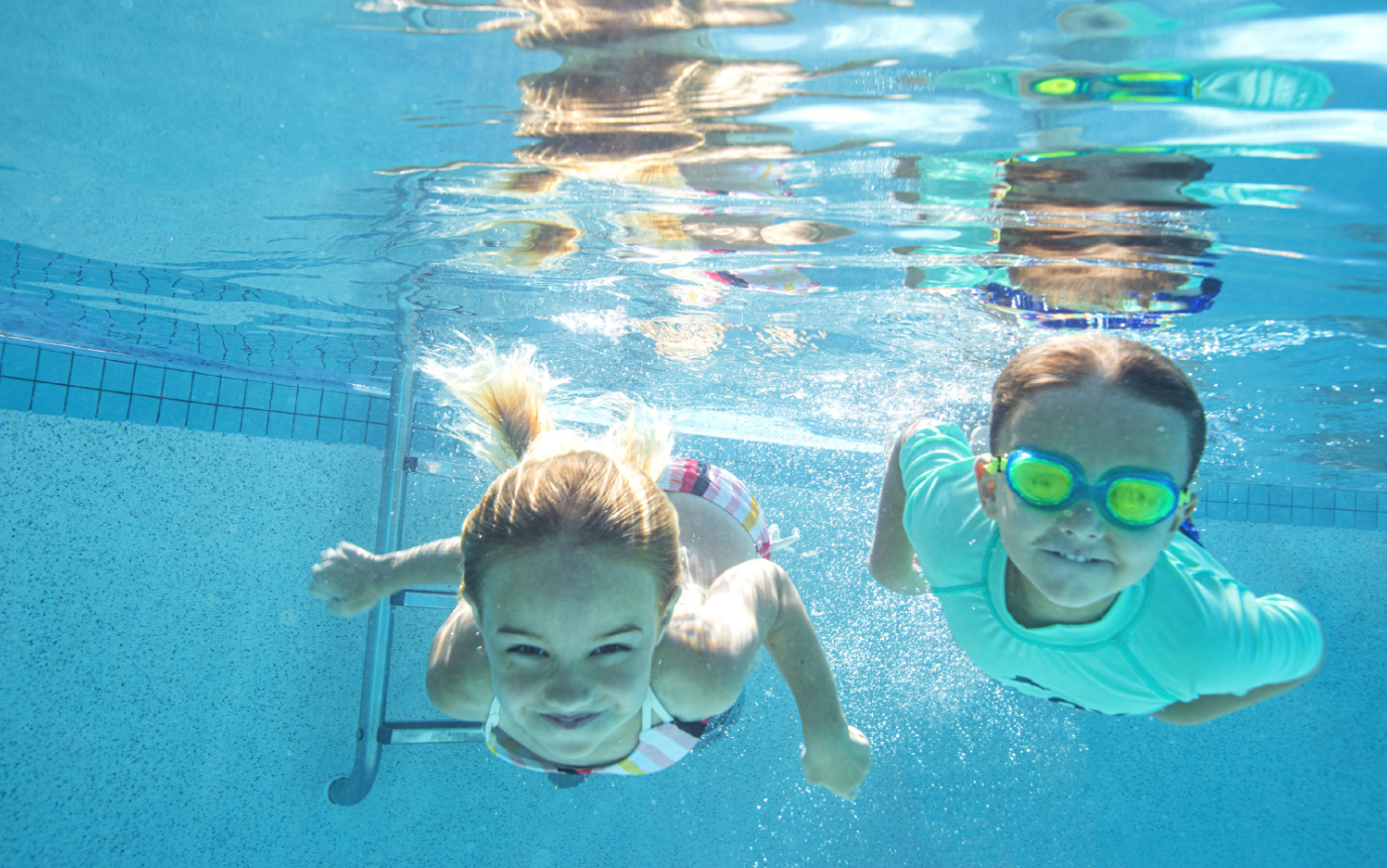 Two children swimming underwater in a pool, smiling, one wearing goggles.