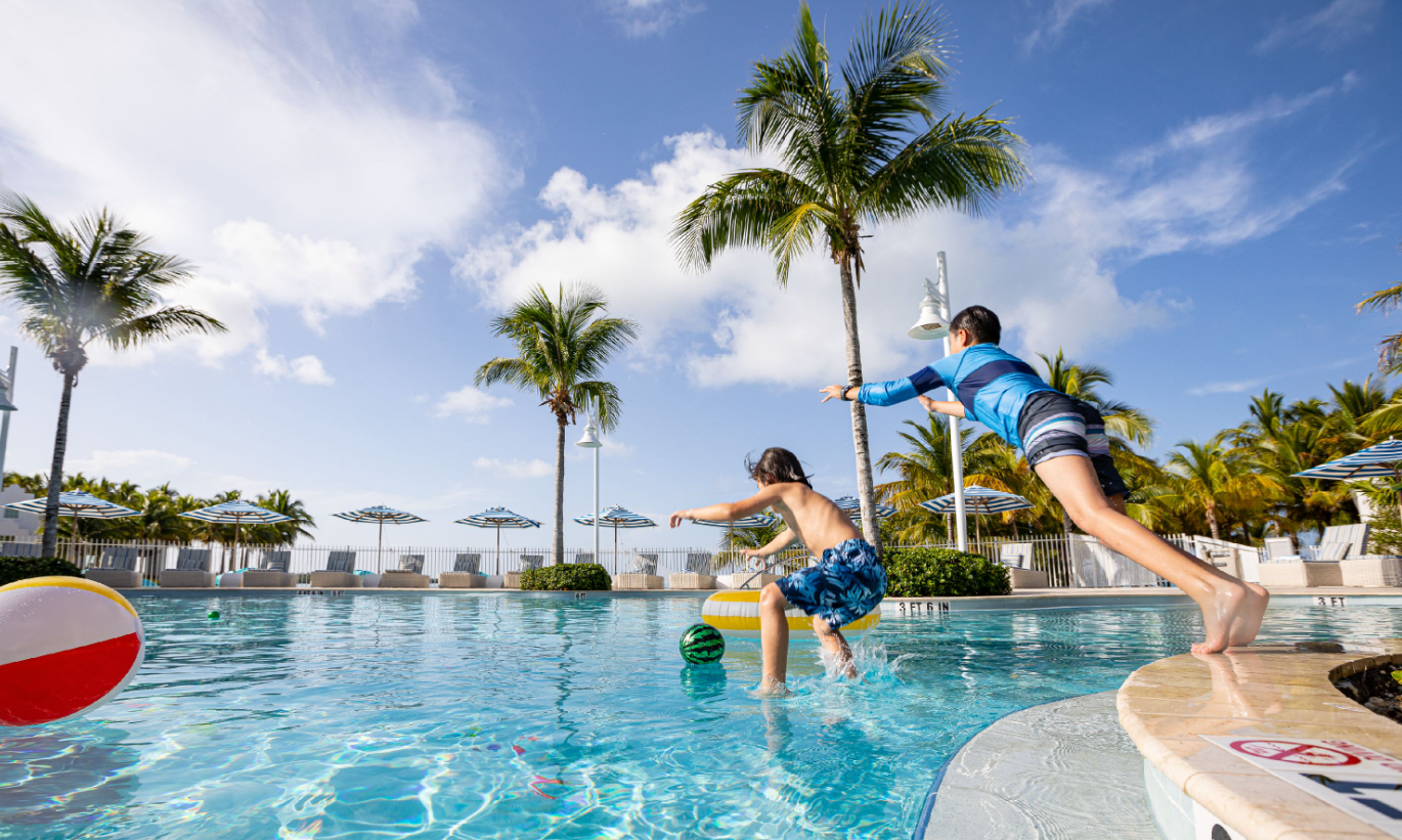 Children jumping into a pool, palm trees and blue sky in the background.
