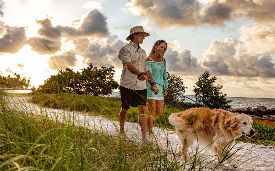 Couple walking a dog on a beach path at sunset, clouds in the sky.