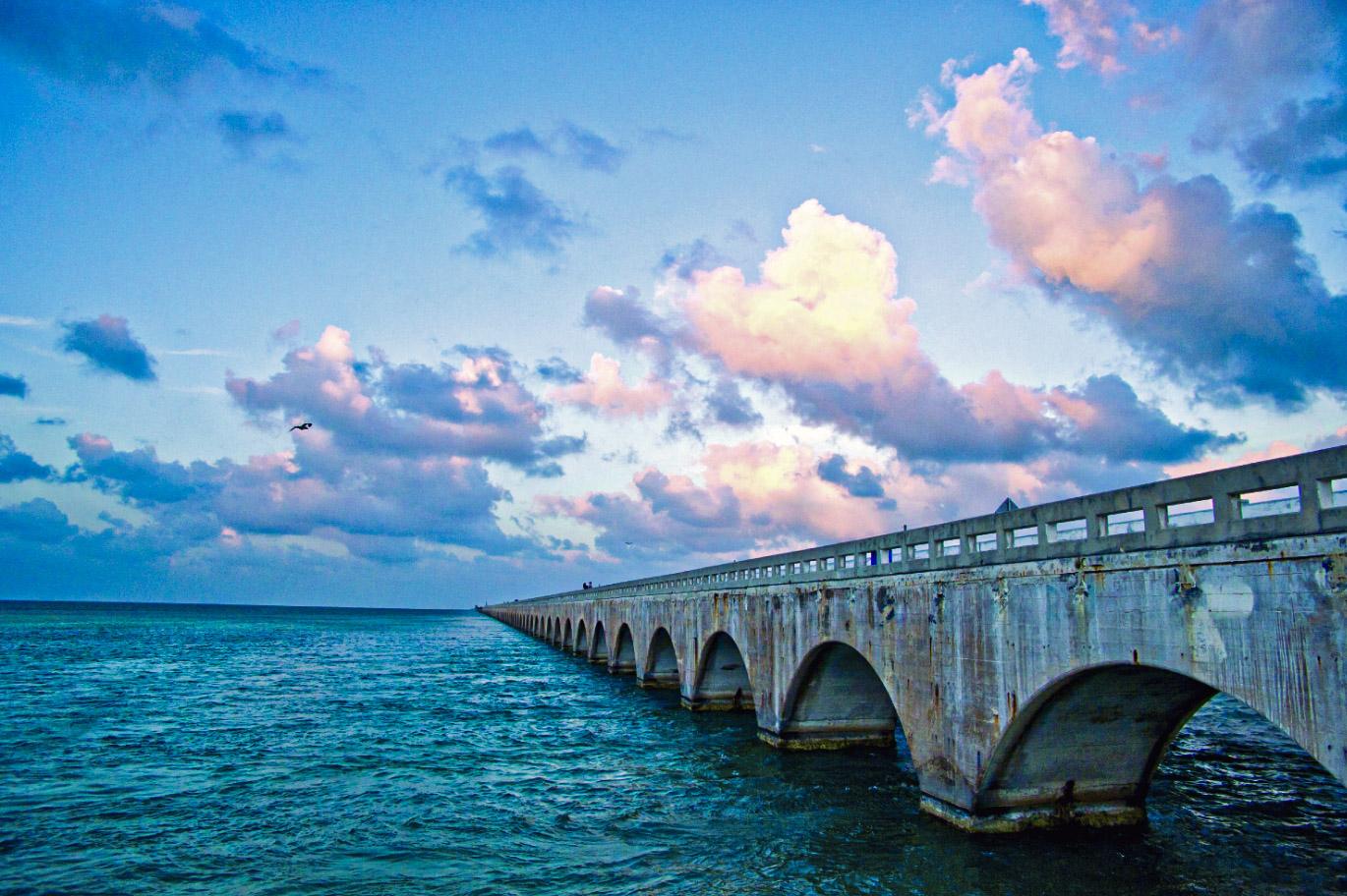 Bridge over ocean at sunset with colorful clouds.
