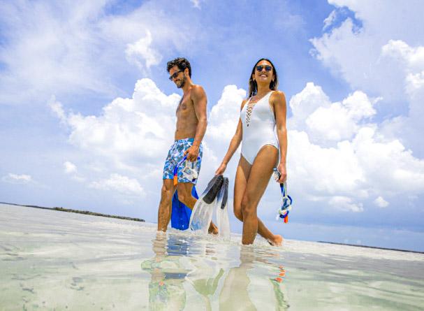 Man and woman walking in shallow water under a bright sky.