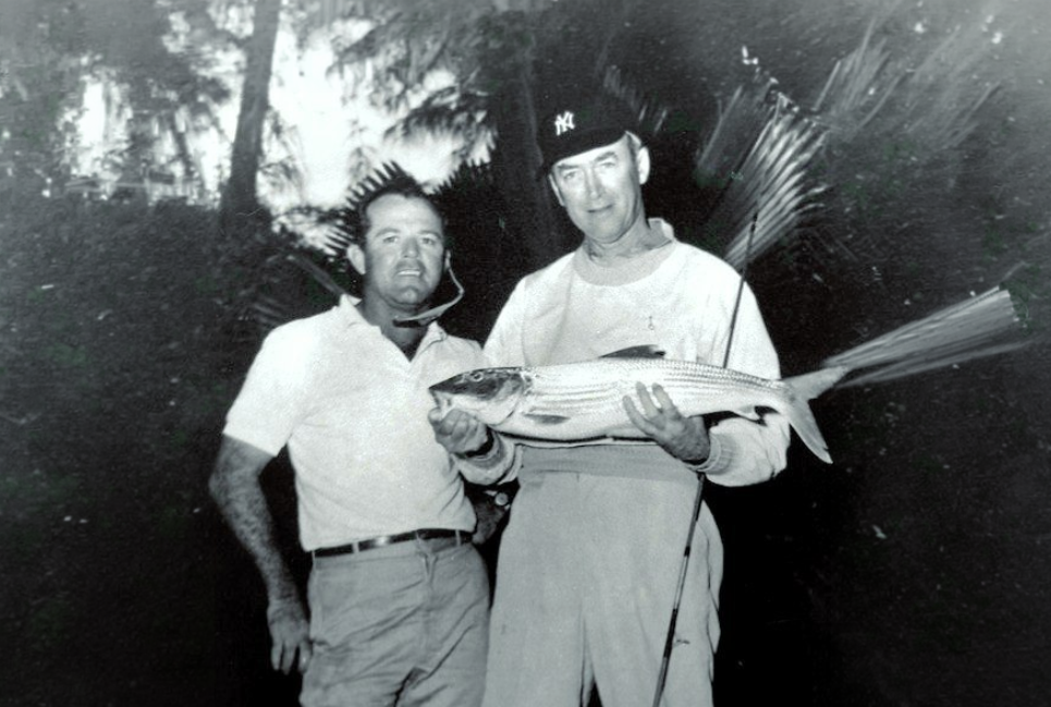 Two men stand together, one holding a large fish, in a black-and-white outdoor setting.
