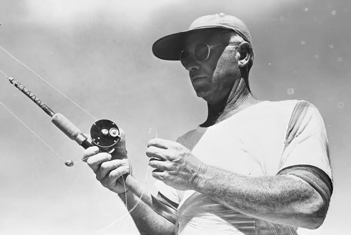 Man holding a fishing rod, wearing a cap and glasses, set against a cloudy sky.