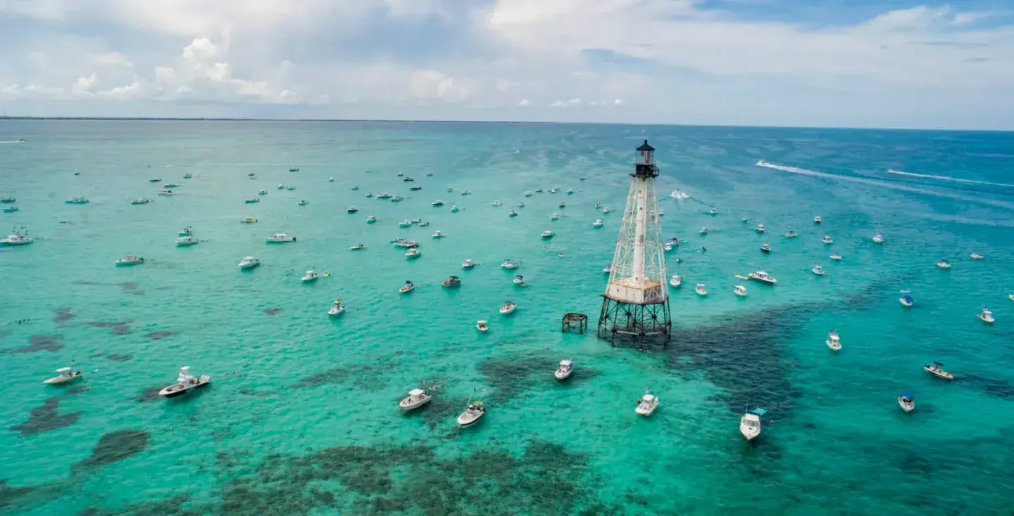 Aerial view of a lighthouse surrounded by boats on turquoise water.