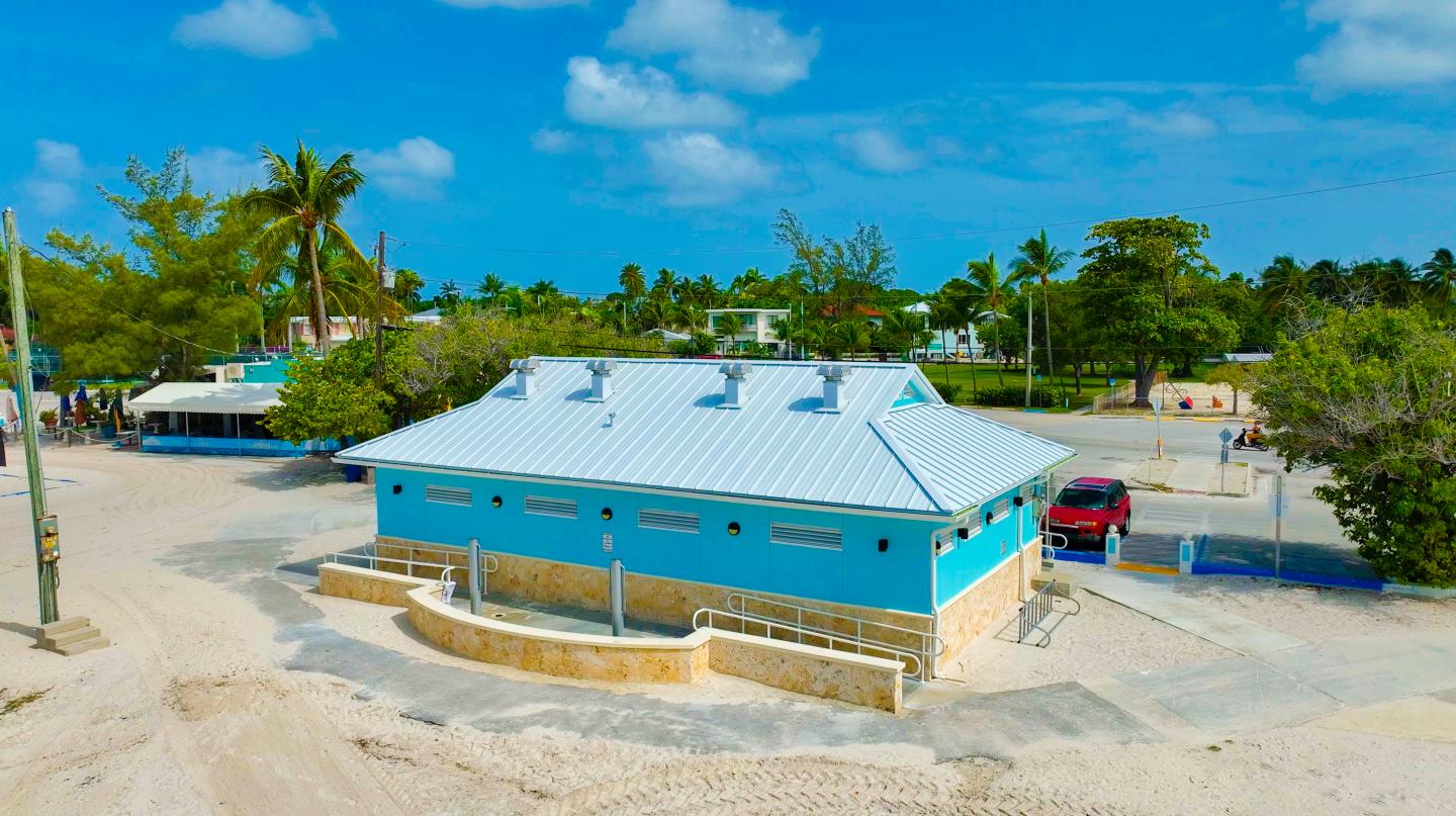 Blue building with a white roof near a beach, surrounded by trees and a parked red vehicle.