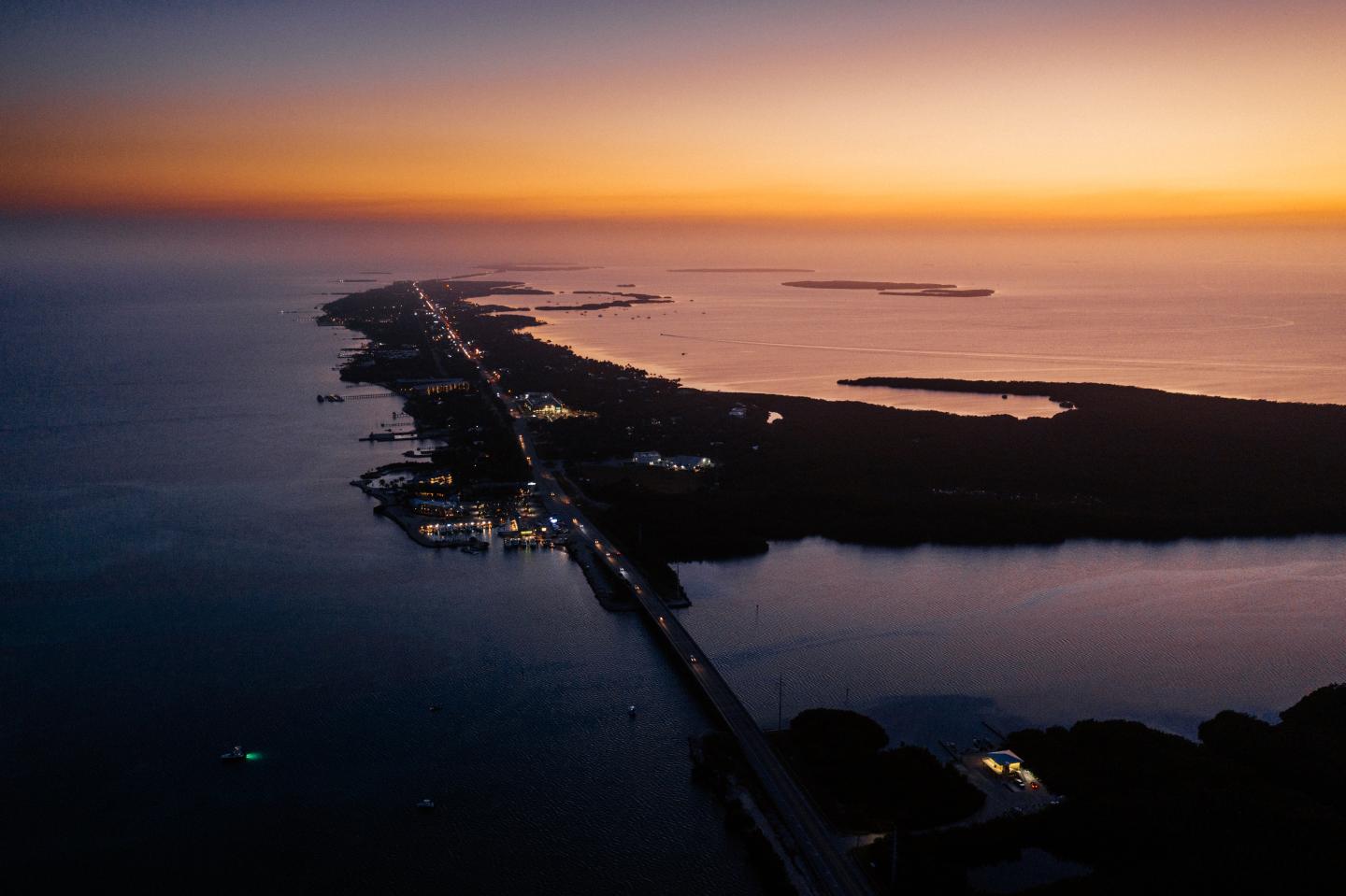 Aerial view of a coastal road at sunset, with dark water and glowing horizon.