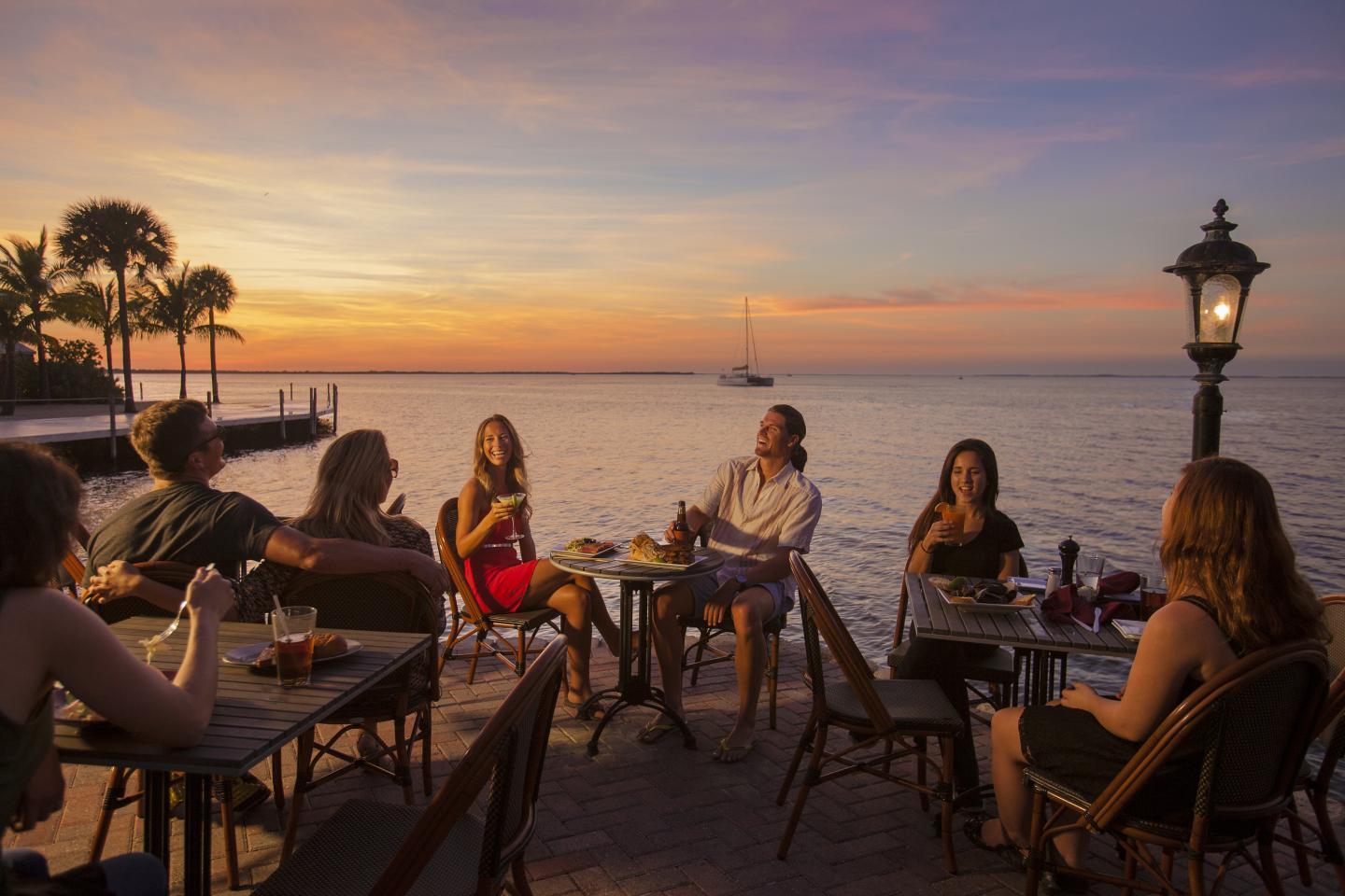 Sunset dining by the sea, friends gather around tables, palm trees in the background.