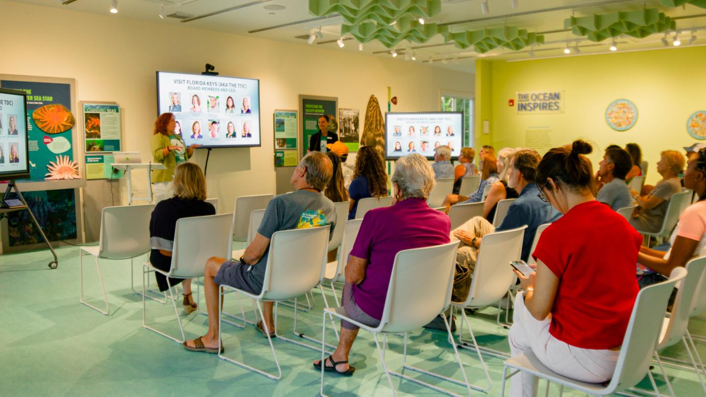 Audience seated in a bright room, watching a presentation on two screens.