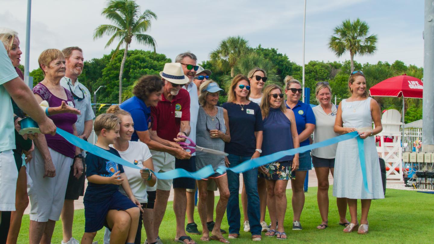 People gathered outdoors cutting a blue ribbon, palm trees in the background.
