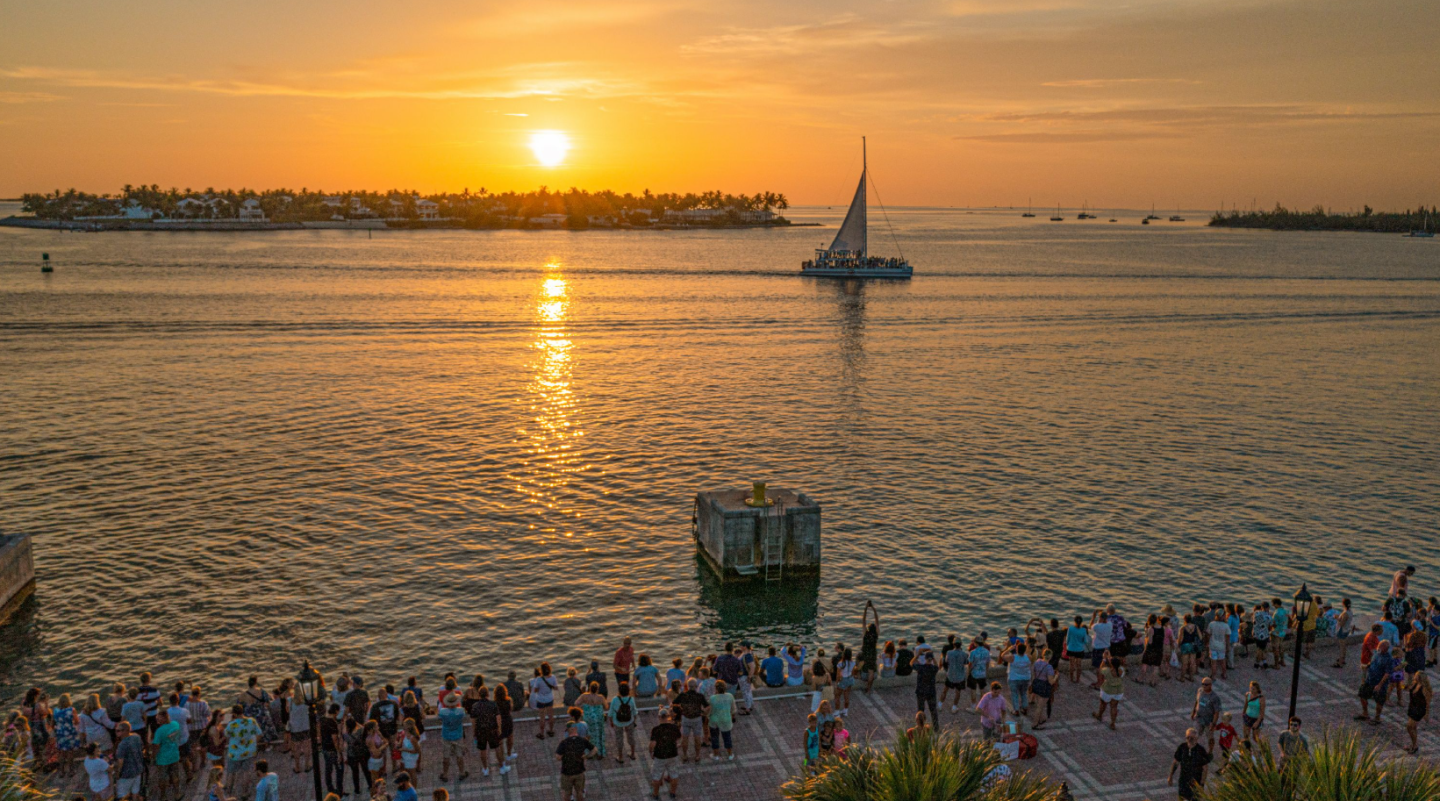 Sunset over water with sailboat; crowd watches from the waterfront.