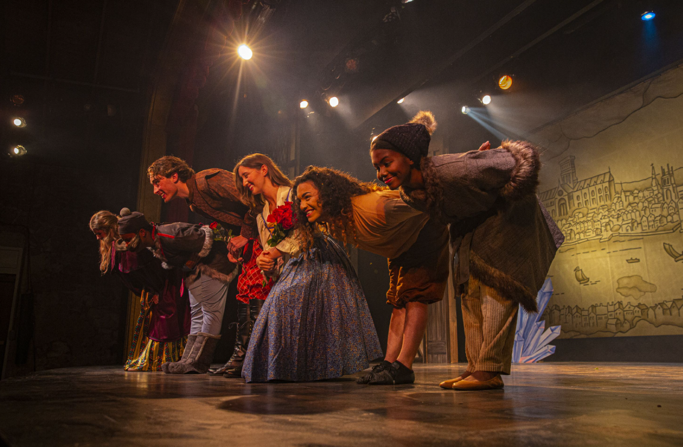 Actors in costume take a bow on stage under warm lights.