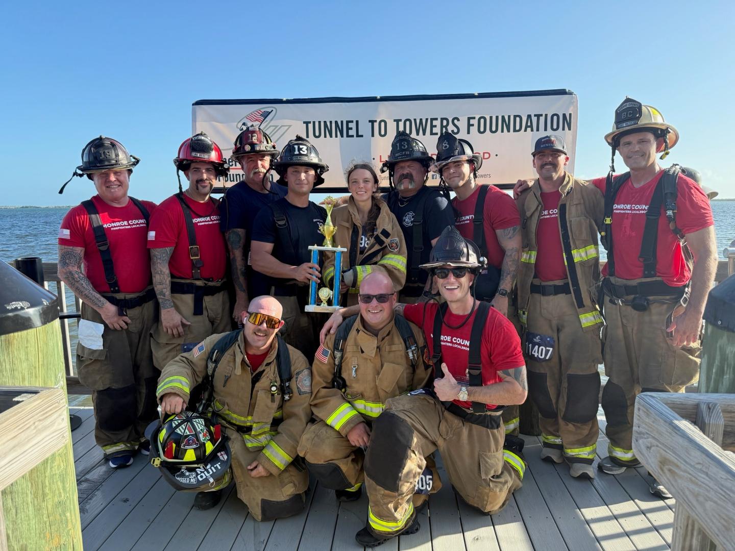 Firefighters in gear pose with a trophy on a dock, clear blue sky above.