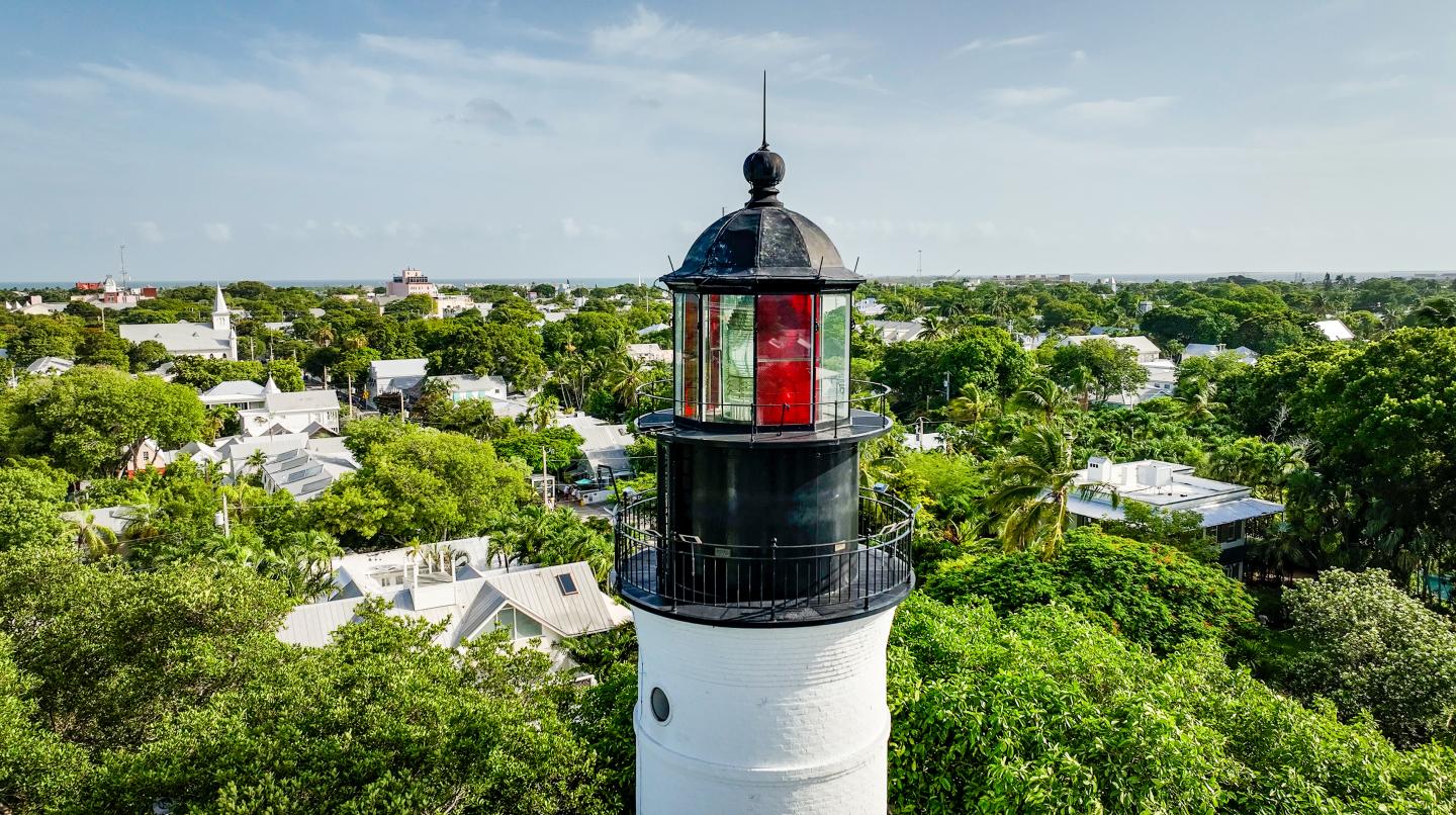 Lighthouse above a tree-covered residential area under a blue sky.