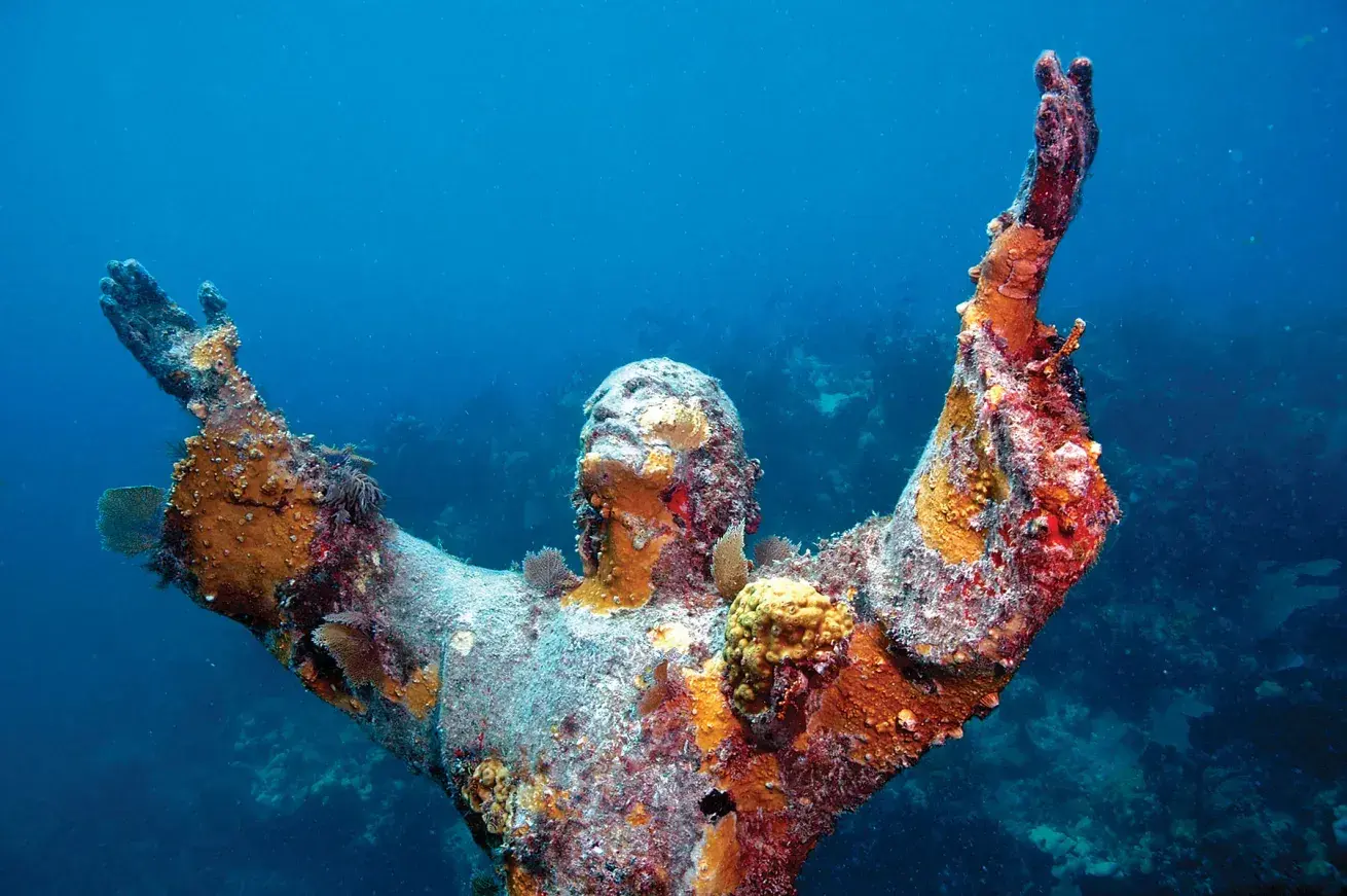 Underwater statue of a man with arms raised, covered in colorful sea growth.