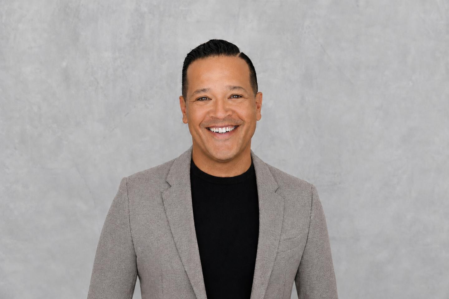 Smiling man in a gray blazer and black shirt against a gray backdrop.