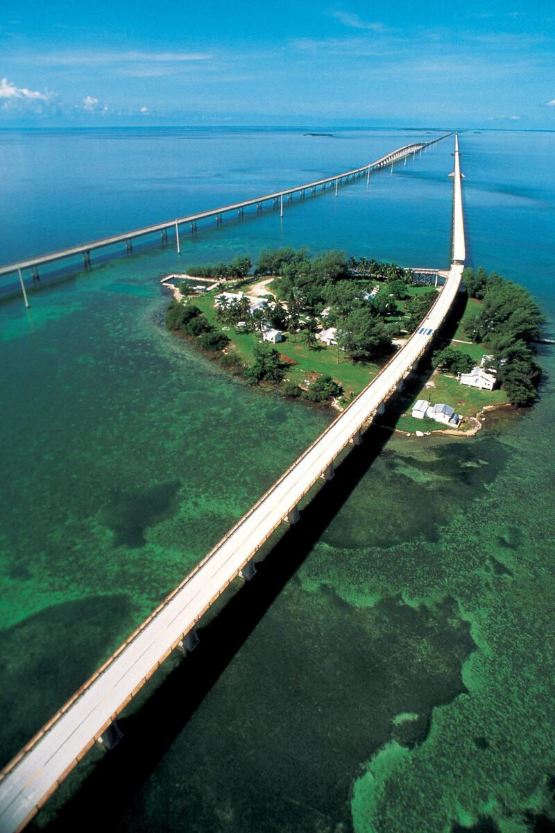Aerial view of a long bridge crossing turquoise water to a small green island.
