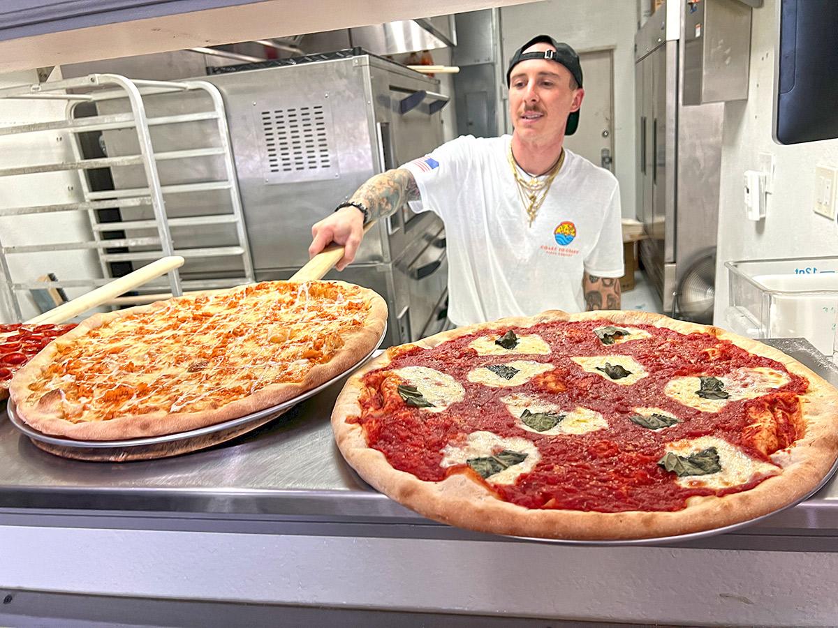 Chef holding a pizza peel behind two large pizzas in a kitchen