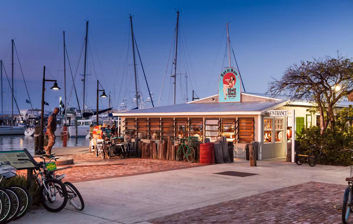 Cyclists outside a warmly lit café at dusk, with sailboat masts behind.