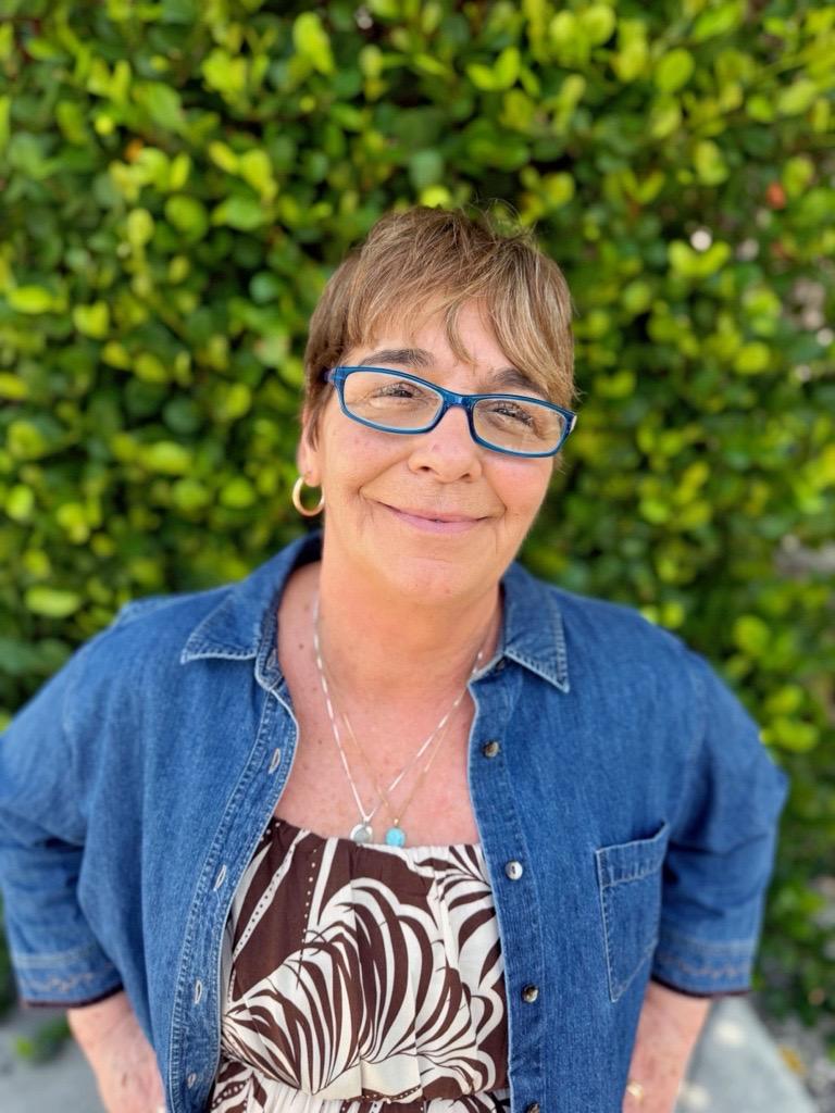 Portrait of a smiling woman in glasses and a denim shirt before green foliage.