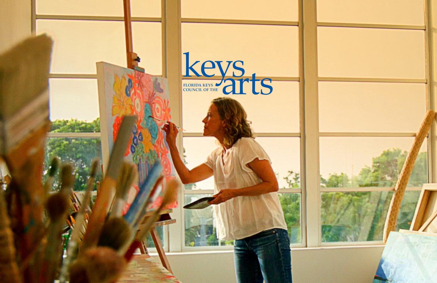 Woman painting on a canvas in a sunlit art studio with large windows.