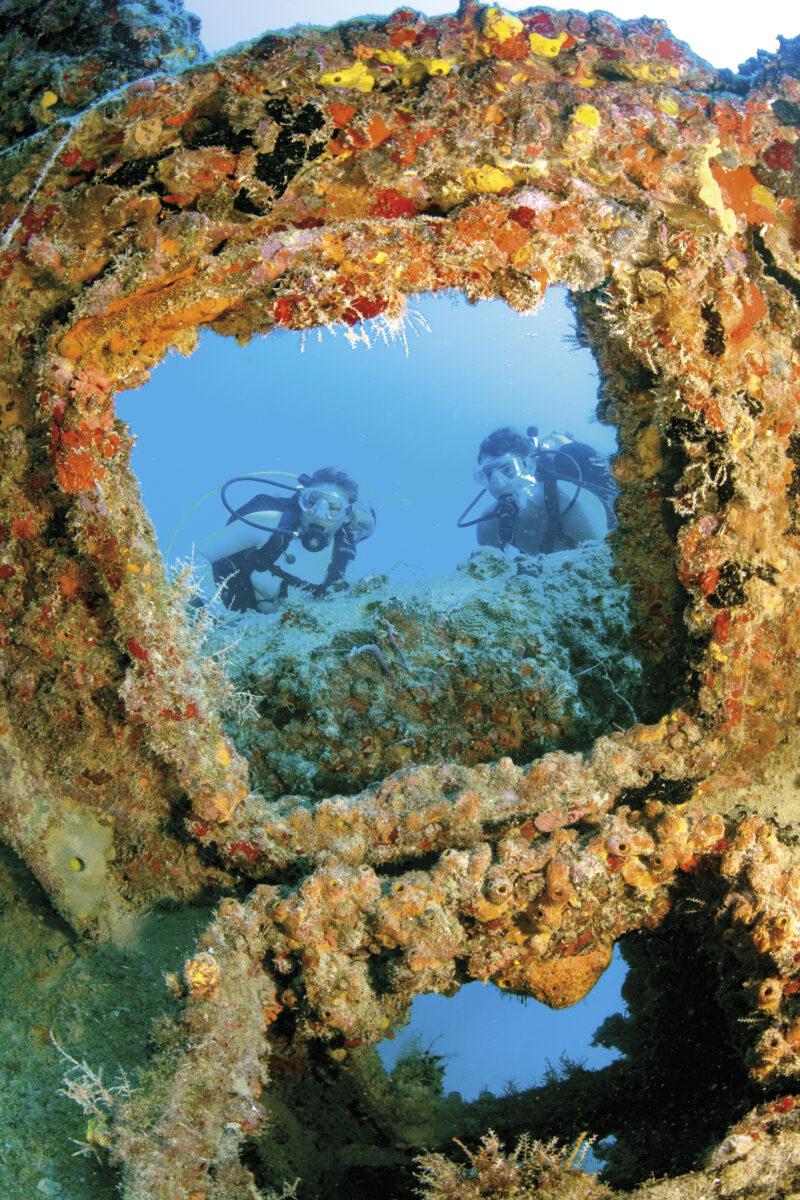 Scuba divers swim through a coral-encrusted shipwreck opening.