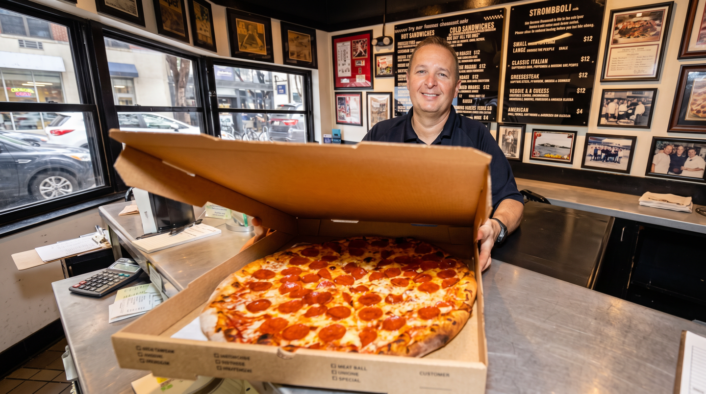 Smiling woman presents a large pepperoni pizza in a restaurant.
