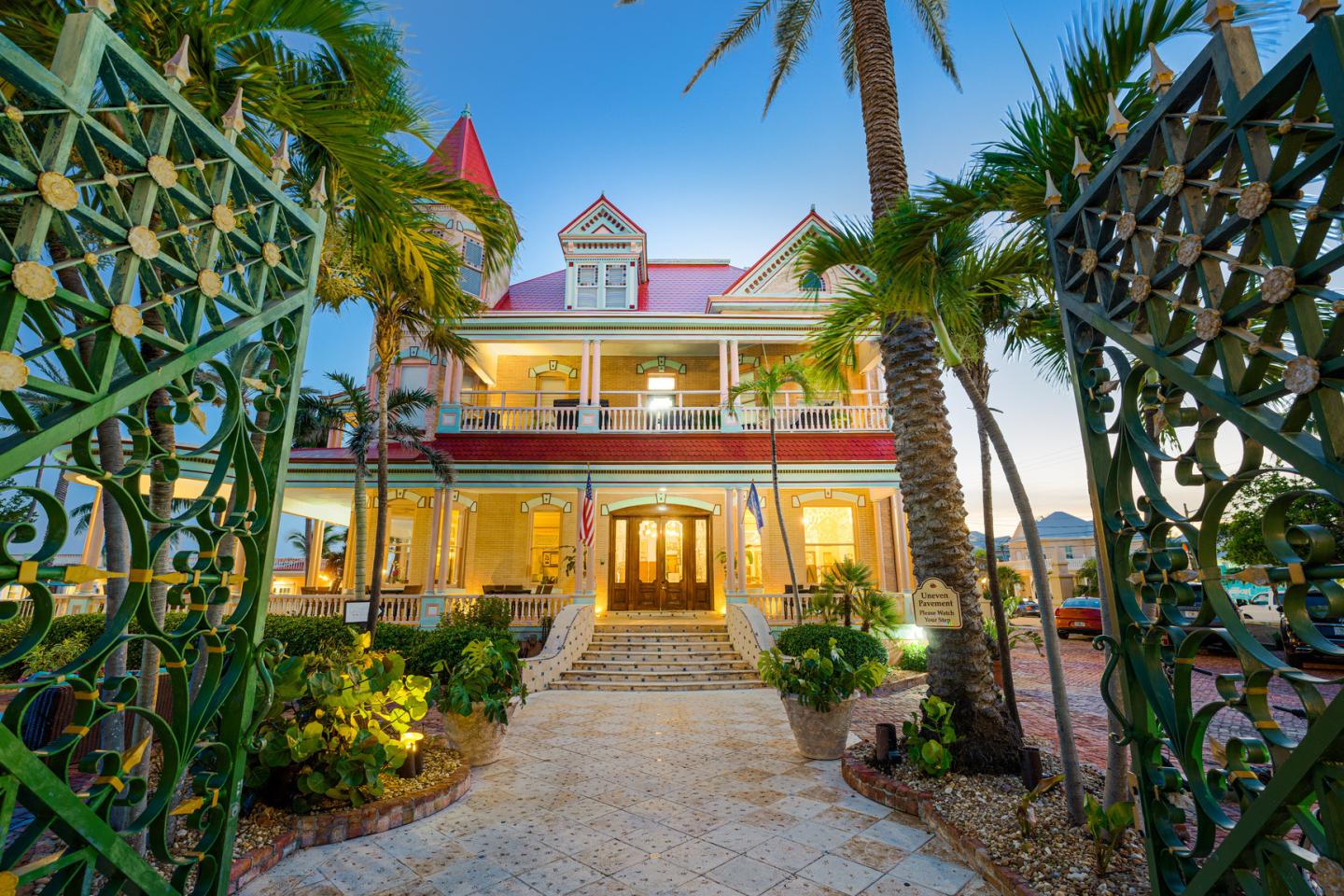 Ornate gates frame a palm-lined path to a brightly lit colonial mansion.