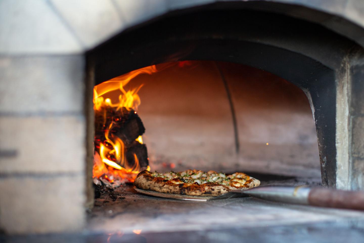 Pizza baking in a wood-fired oven with flames beside it.