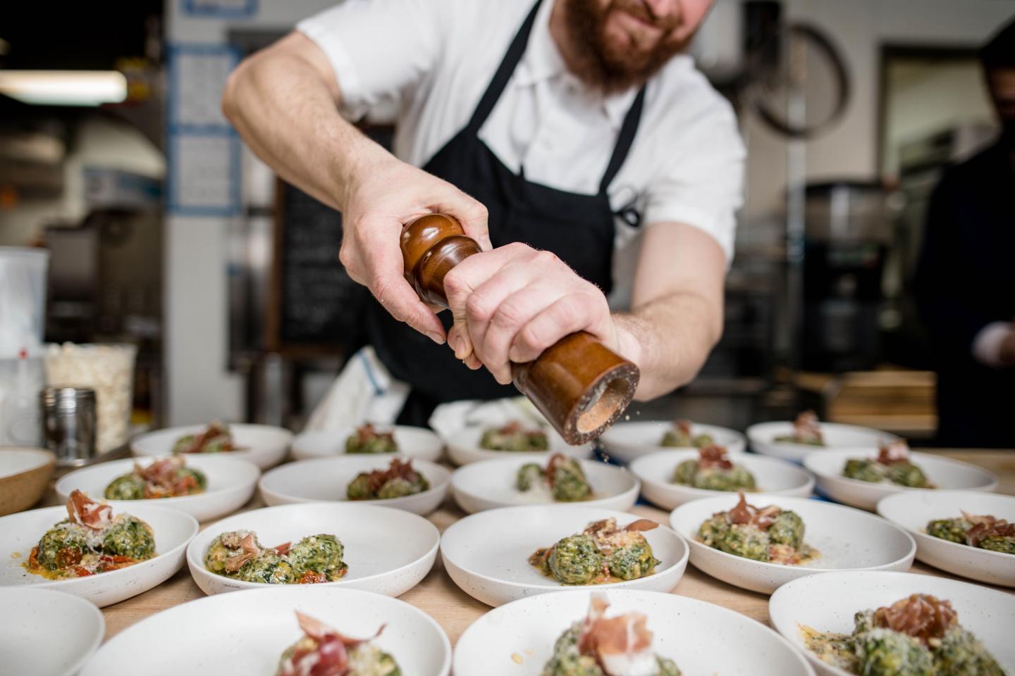 Chef drizzles sauce over plated dishes in a busy restaurant kitchen.
