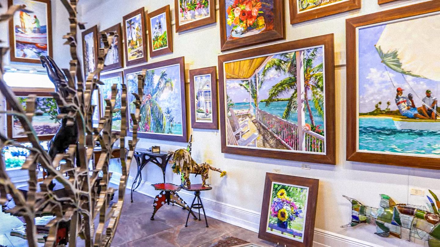 Art-filled gallery interior with framed paintings, sculptures, and a bicycle.