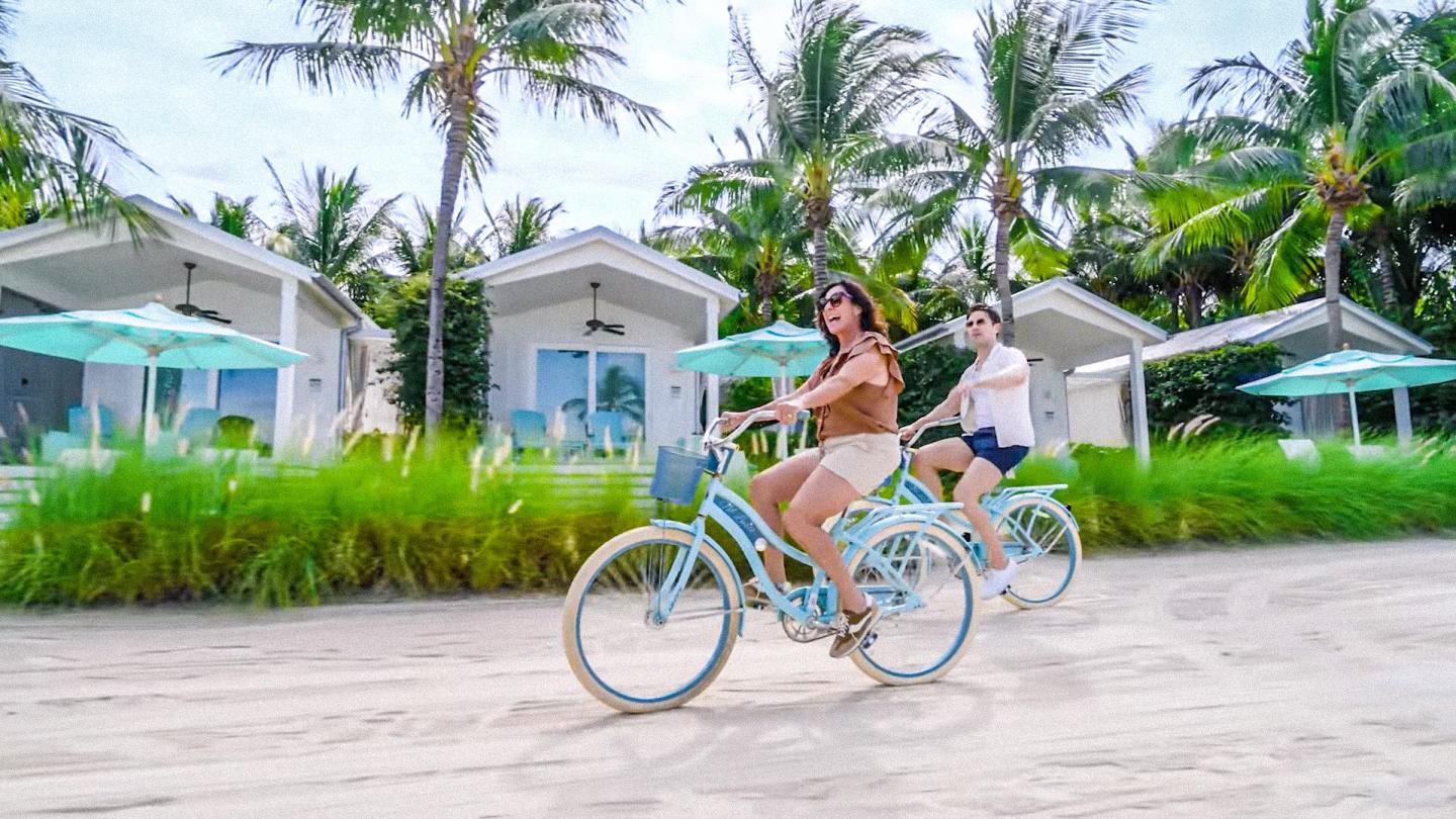 Woman riding a turquoise bicycle past tropical beach cottages and palm trees.