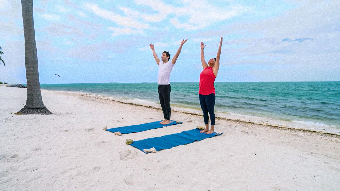 Two women practicing yoga on blue mats on a beach by turquoise water.