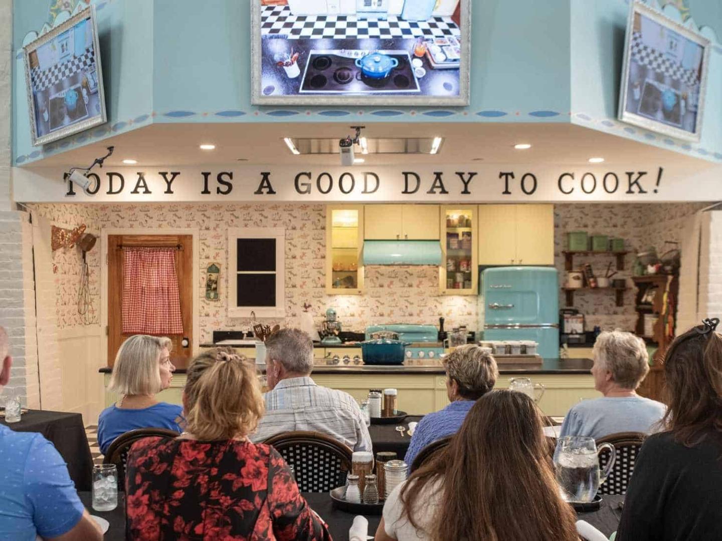 Cooking demonstration in a cozy kitchen before a seated audience.