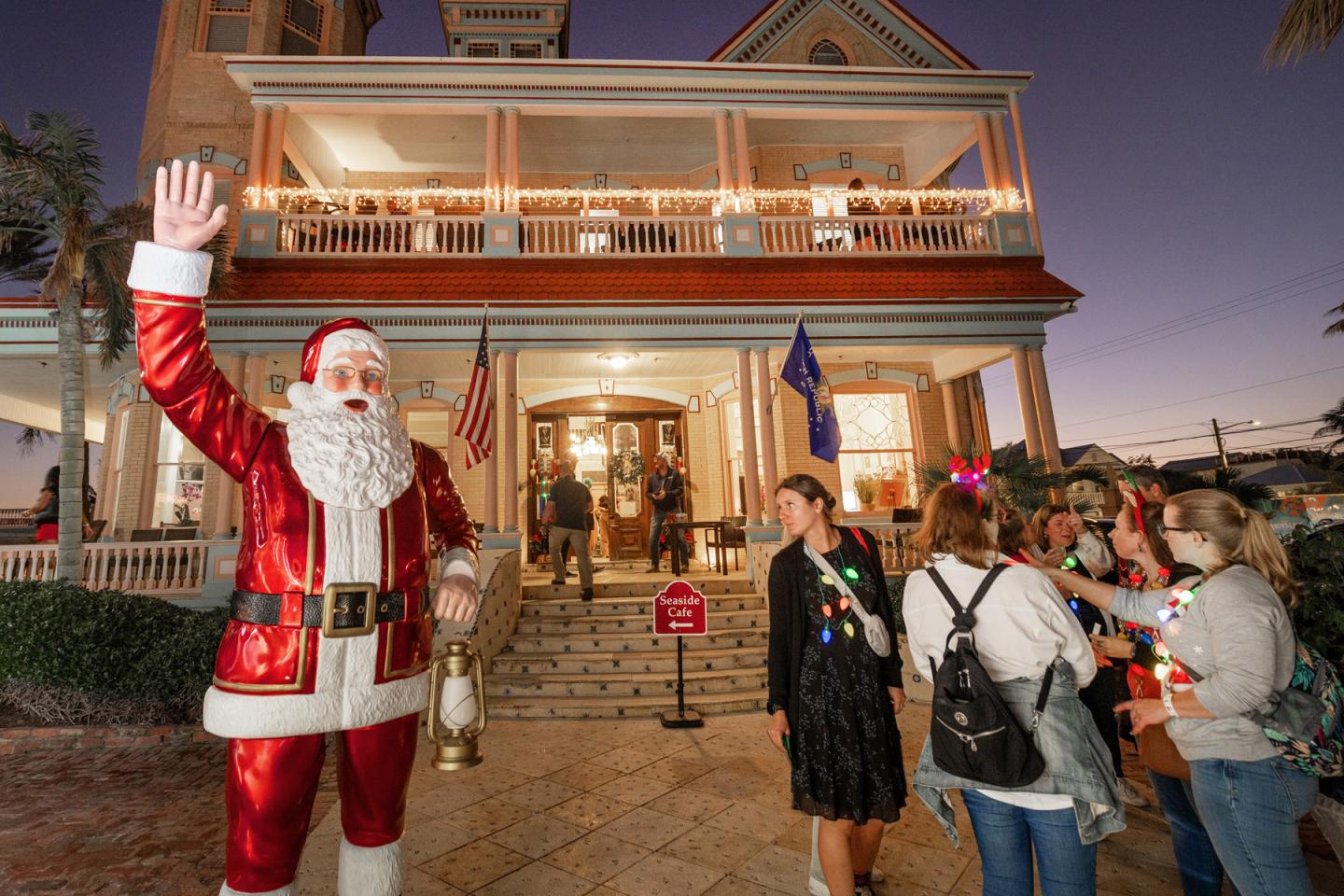 Santa waves outside a festively lit Victorian house as visitors gather.